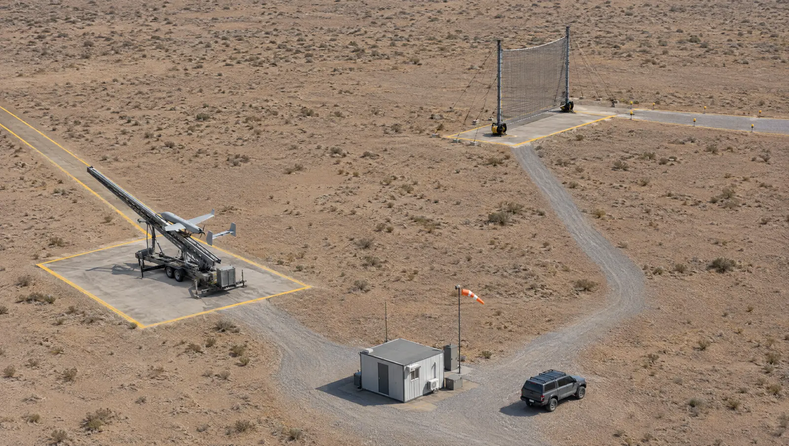Aerial view of a launch and recovery range: a trailer-mounted rail launcher aimed down a marked launch corridor, and a separate guyed two-mast recovery net about 150 m away with its own marked approach lane at right angles to the launch direction
